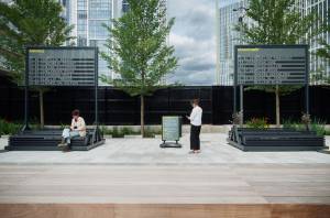 Two boards, one 'arrivals' and one 'departures' are situated in the Nine Elms, Embassy Gardens courtyard showing a list of names and dates. One person stood in between the boards and one person sat in front of a board