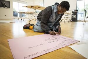 Anna Maria Nabirye, a black woman with short hair and a denim outfit, smiles as she writes on a big piece of paper