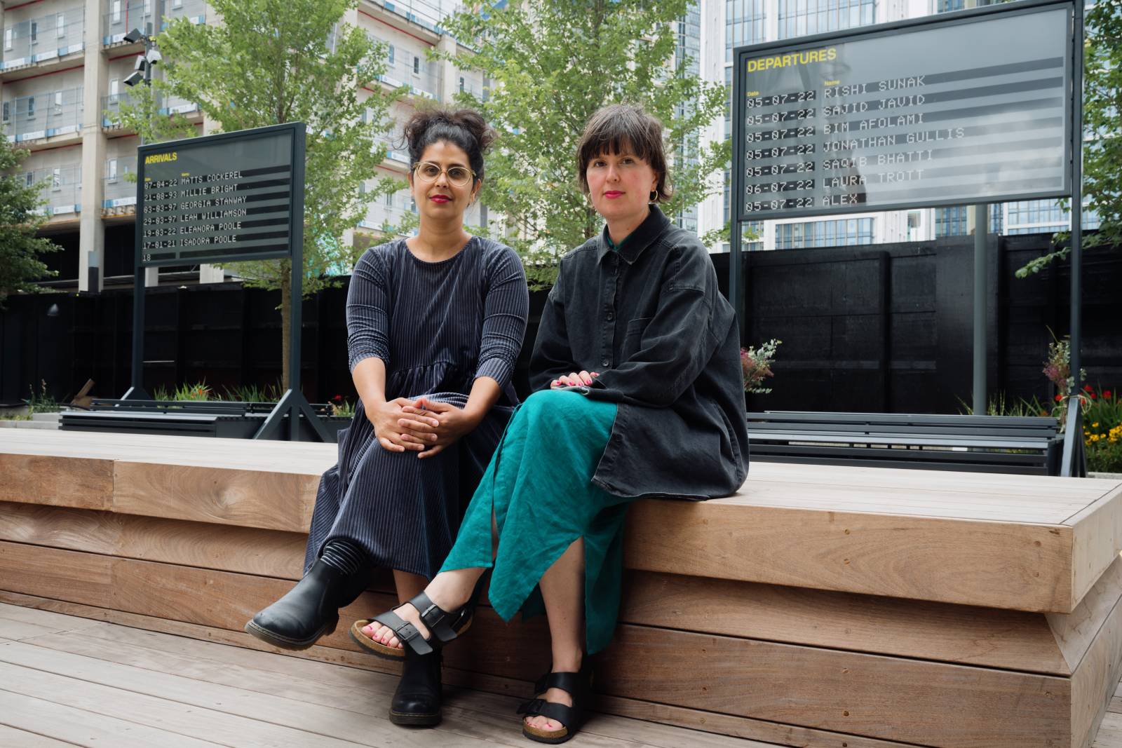 YARA + DAVINA sat on a wooden bench in front of arrivals and departures boards at a courtyard in Nine elms, Embassy Garden