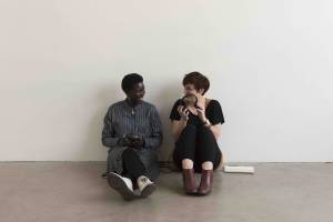 Anna Maria Nabirye and Annie Saunders, a black woman and a white woman, both with short hair, sit against a white wall having a conversation