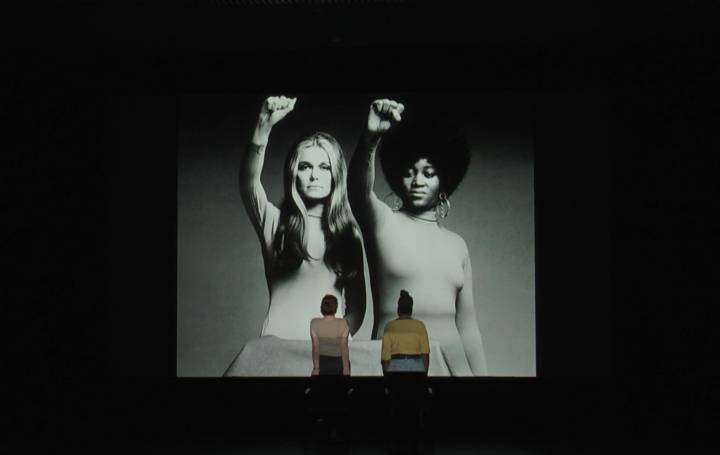 Anna Maria Nabirye and Annie Saunders, a black woman and a white woman, stand facing a large projection of a black and white image ofDorothy Pitman Hughes and Gloria Steinem, taken by Dan Wynn