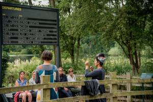 YARA + DAVINA speak to a crowd in front of one of the Arrivals + Departures boards, by the lake in Yorkshire Sculpture Park