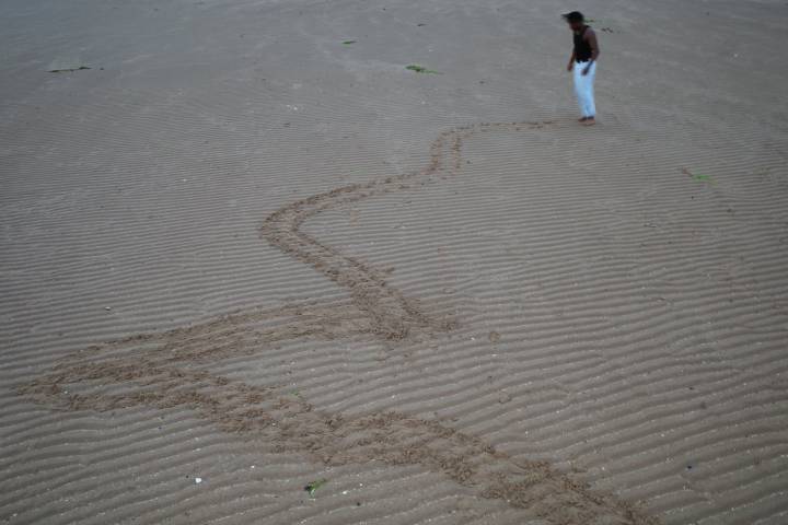 A photo of a figure walking along sand, leaving a trail behind them