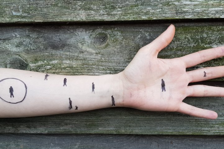Person's arm stretched out against wooden fence background. There are pen drawings of stick people walking across their arm.
