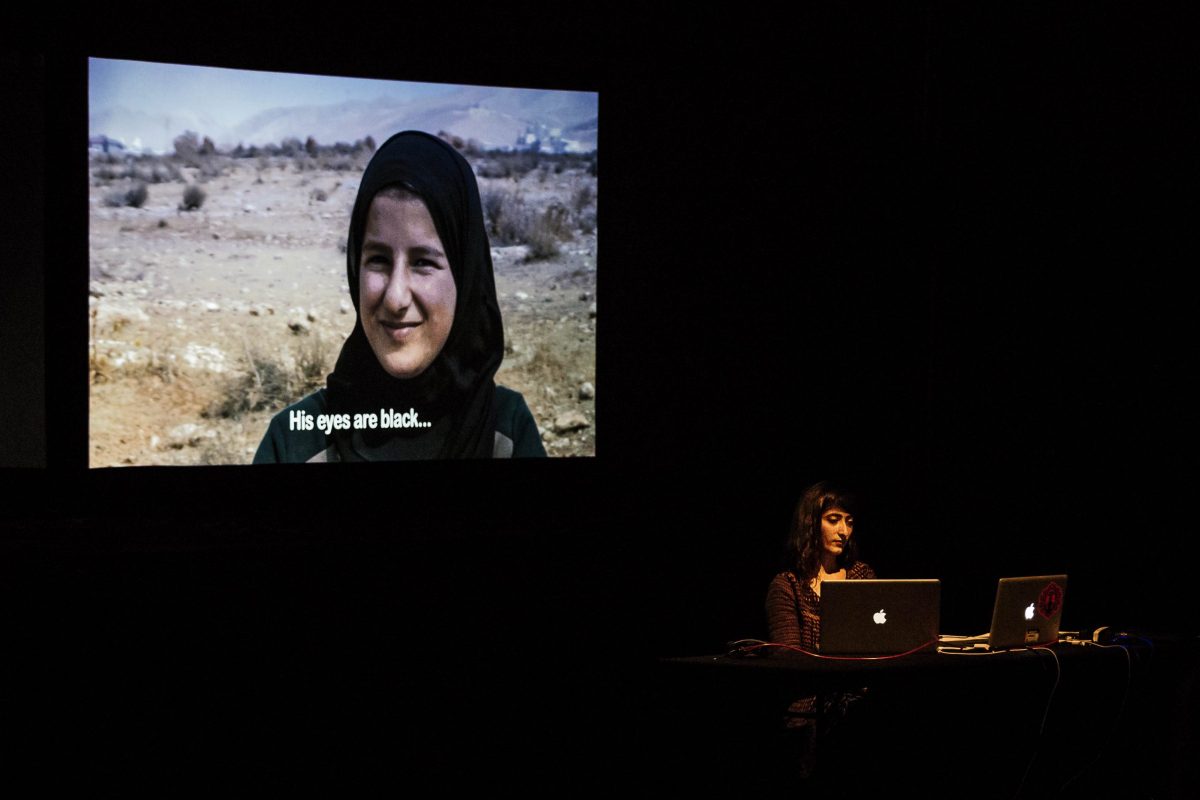 Umama is sat on a stage, in a dark set with a large projection behind her of a clip of a woman being interviewed in a desert