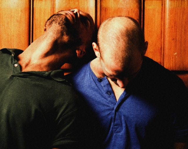 Close up shot of two people stood side by side leaning on each other. The person on the left's head is leant back on the other persons shoulder and the person on the right has their head tilted forward to the floor