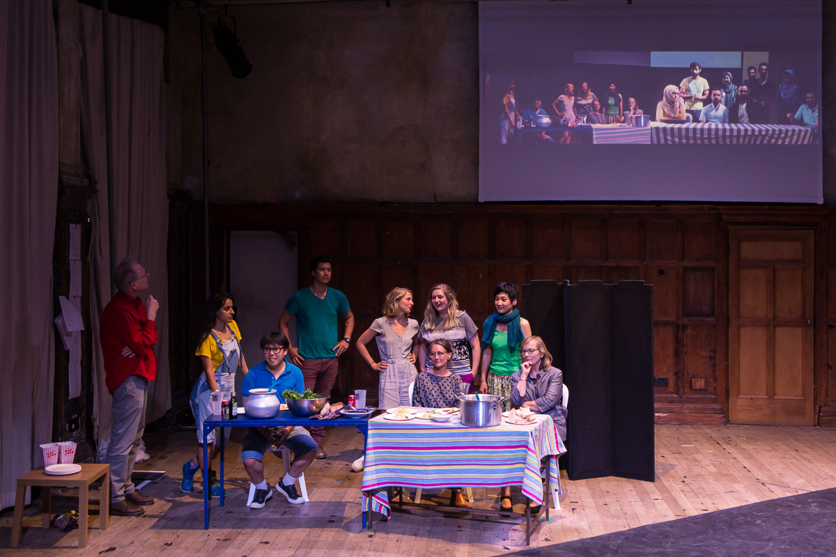 10 people on a stage, 5 gathered around a table, 2 cooking food, a projection of a similar scene in Gaza is on the screen behind