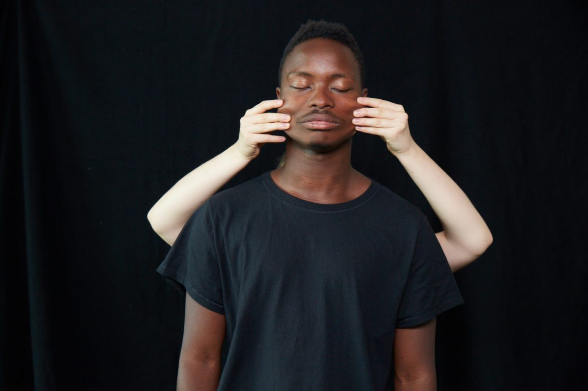 Two people standing with black background. one person standing behind another with hands out pulling their face.