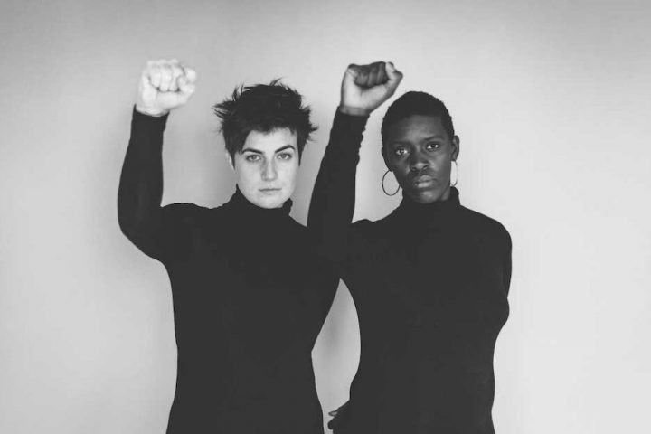 Two people holding their right arm in the air, facing the camera, wearing dark turtle necks against a white backdrop. A black and white photograph