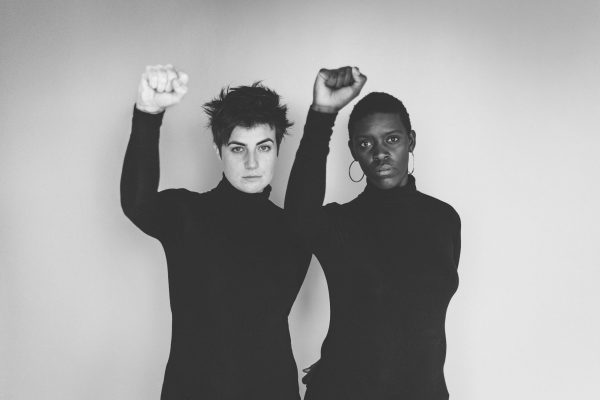 Two people holding their right arm in the air, facing the camera, wearing dark turtle necks against a white backdrop. It is a black and white photograph