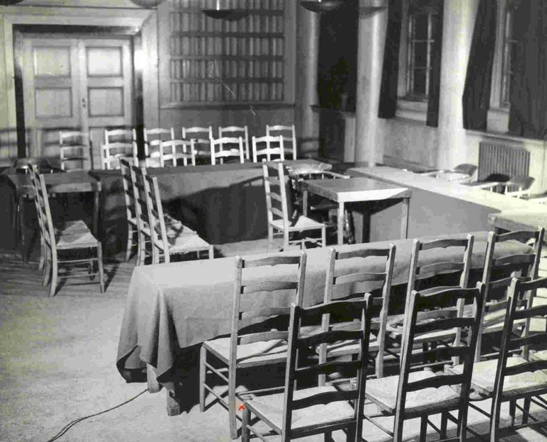 Black and white photo of room with wood panelled walls, tables and chairs