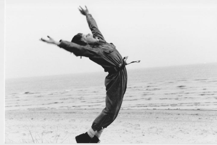 A black and white image of a young boy on a beach leaping with joy with his arms in the air. He is wearing a dark boiler suit with a belt adorned with shells.