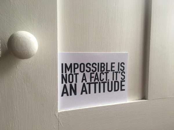 closer up of white door with handle on left and postcard sitting on a ledge stating "impossible is not a fact, it is an attitude"