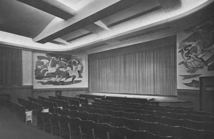 Black and white photo of a theatre with rows of seats, a stage with curtain down and murals on either side of the stage