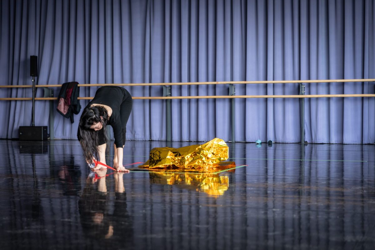 Performer in dance studio putting tape on the floor. There is a gold foil blanket on the floor next to them.