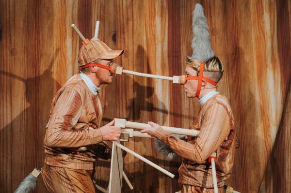 There are two people facing each other against a fake wooden backdrop, wearing fake wooden outfits with real wooden appendages, which are joined at the nose