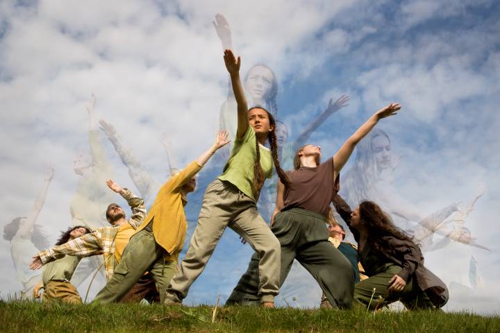 A photo of an intergenerational group of people performing in dynamic, powerful postures with grass below and blue sky above
