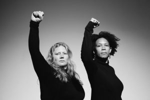 Two people holding their right arm in the air, facing the camera, wearing dark turtle necks against a white backdrop. It is a black and white photograph