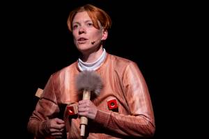 Rosana Cade, a non-binary person with short hair, looks into the audience from a dark stage. Rosana wears a fake wooden outfit and holds a microphone