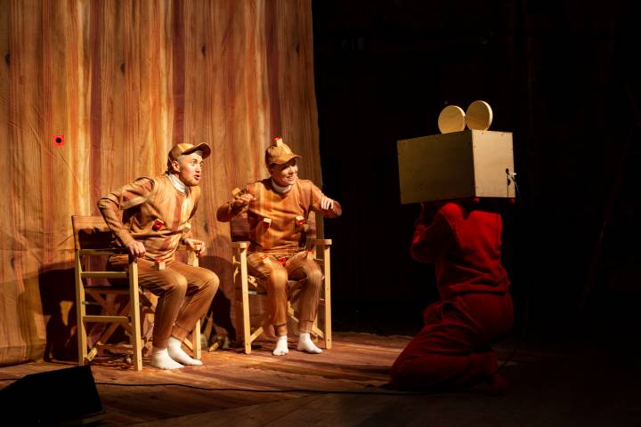 Two people in fake wooden outfits, in a film studio setup made of wood, sit on deck chairs and face a person with a camera for a head