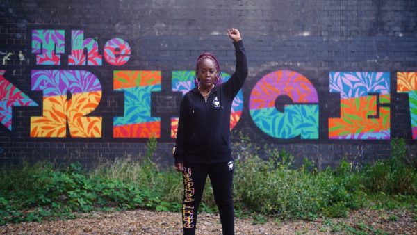 Lady Unchained stands in front of a graffitied wall with right fist raised in the air