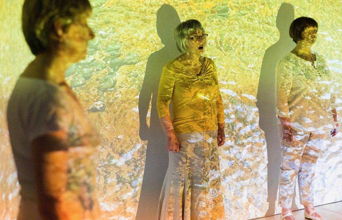 Three people dressed in white, standing and singing with yellow abstract projections on them