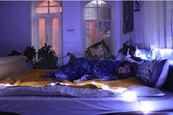 Person lying on a bed in a half-lit bedroom. They are wearing a dressing gown, lying on top of a blanket. There are plants, a large window and a door in the background.