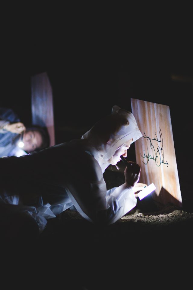 Person in white protective suit lying in a dark place on the soil, holding a torch to read a card. another person is lying behind them, ear to the ground