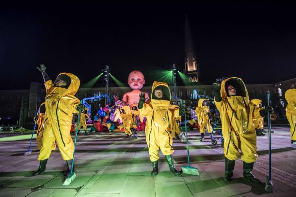 Three people in yellow hazmat suits hold brooms, gazing outward. It is night time and they are standing on a square somewhere. Behind them is a big white inflatable baby, more people in yellow hazmat suits, and a circus playground. Two tall pillars beam out green light onto the scene.