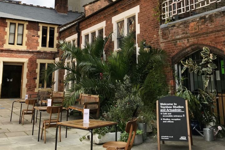 Outside of a red brick building with palm trees, tables and chairs on a courtyard