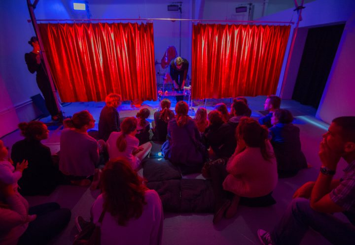 Seated audience of around 20 people watching a performer on stage arranging objects on a table. There are red curtains in the background on railings