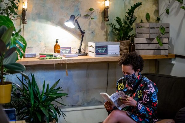 person sitting on an armchair reading a book, wearing a mask. They are surrounded by plants, books and lamps
