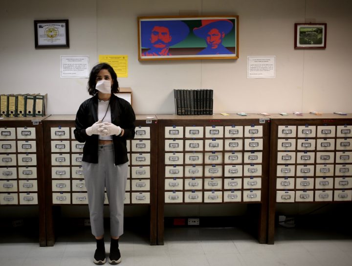 A woman stands in front of a series of small boxes. She is wearing a white facemask and wearing white cotton gloves.