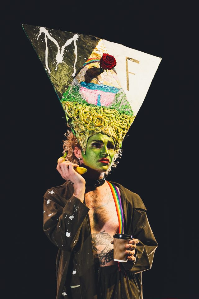 A person with green painted face with red lipstick a large cone shaped painted headpiece, holding banana to ear and coffee cup in other hand