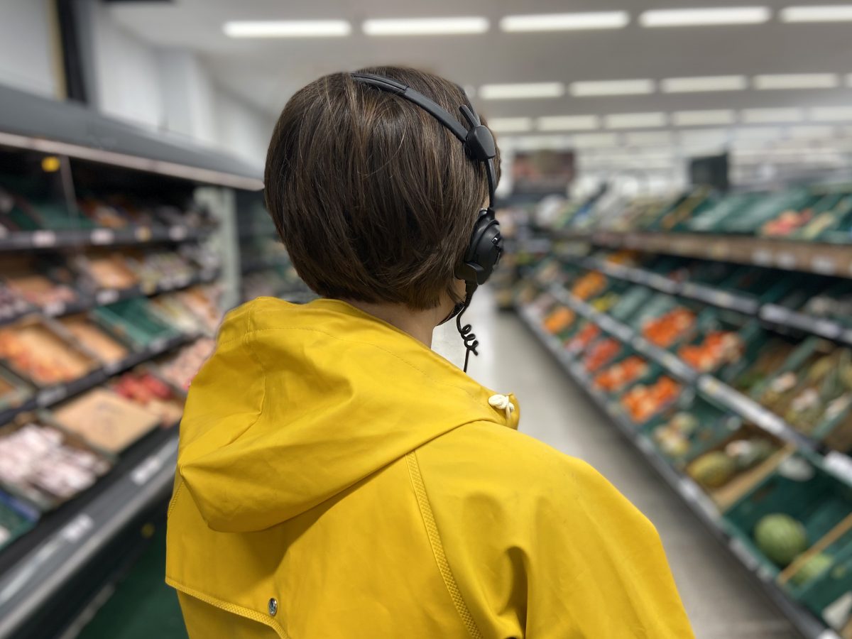 A person with a bob hair wearing a yellow jacket and headphone stands in a super market aisle. They are turned away from the camera and looking at the fruit and veg
