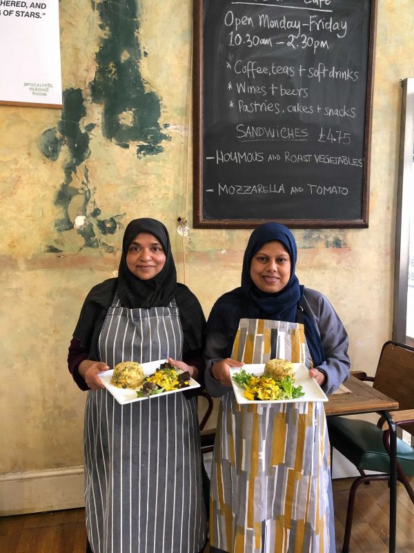 Two women from Tati Canteen in a cafe wearing aprons and holding plates of food