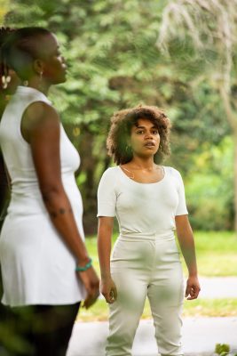 person standing in a park, wearing a white top and trousers with a curly brown afro