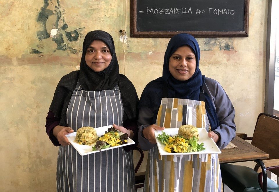 Two women from Tati Canteen in a cafe wearing aprons and holding plates of food