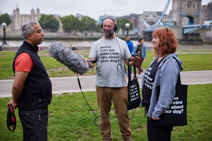 Two people in a park with microphone and recording equipment interviewing a person. The intervewees are wearing 'can I talk to you about your dog' graphic t-shirts