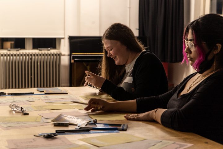 two young people sat next to each other at a table making zines and smiling