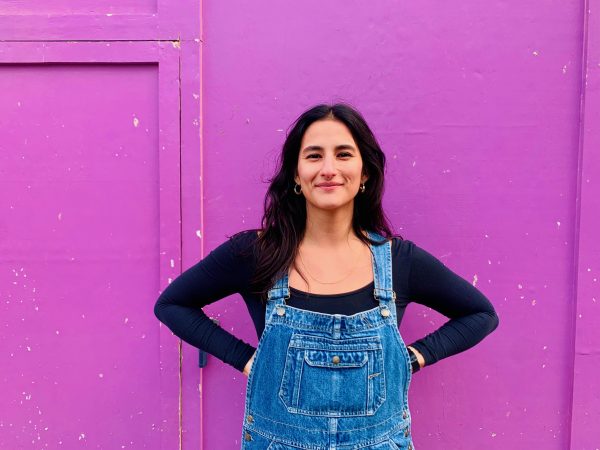 Valentina standing smiling in front a bright purple wall. she has long dark hair and is wearing dungarees.