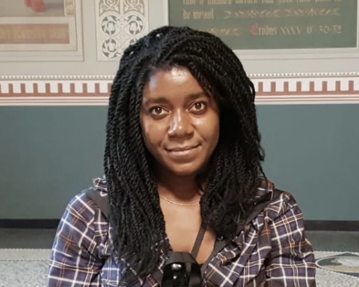 Image of Paige Ofosu-Asaa, a black woman with short twist braids, wearing a plaid shirt and a camera around her neck, smiling into the camera
