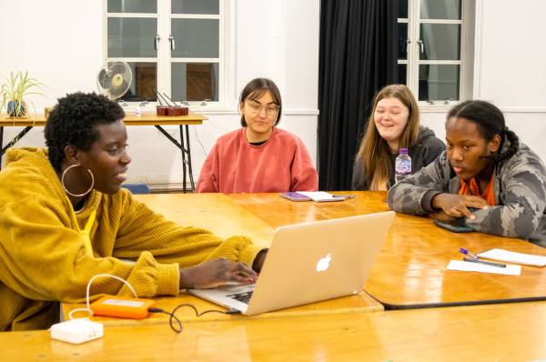 Group of three young people sat around a table laughing and observing content on a laptop with Anna Maria Nabirye