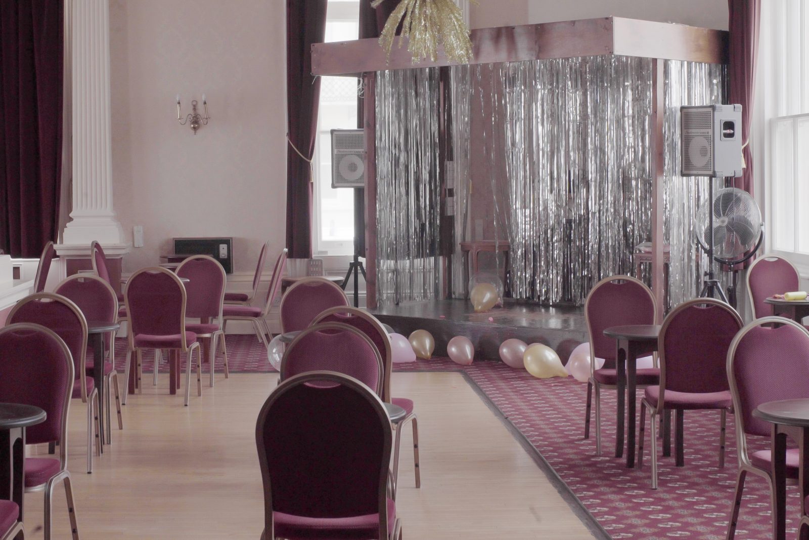 Room in hotel with pink chairs dotted around. There is a silvery foil curtain in the background with pink balloons and a pink carpet at the bottom