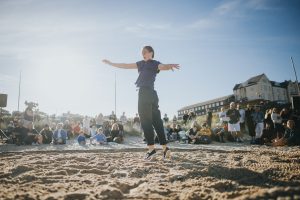 person in a mid spin dancing inside a circle shaped rope on sandy shore with an audience