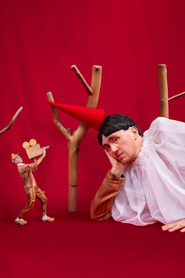 There are two people facing the camera in a rich red backdrop with fake trees made of wood. On the right, the person, Ivor, is lying down, leaning on his elbow. He wearing pointed red hat, a large collar and a top printed with a fake wood pattern. There is a small person on the left, Rosana, and they are wearing a cap, top and trousers printed with a fake wood pattern, with wooden clogs. They are carrying a wooden video camera, pointed at Ivor.