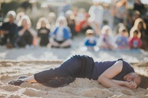 person mid-choreography, lying on the sand inside a circle shaped rope on a sandy shore with an audience