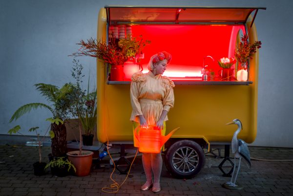 Jennie Moran, stood in front of a yellow food cart with a red neon glow inside holding an illuminated. Jennie is wearing a yellow dress and holding an illuminated red watering can