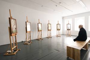 A landscape image of the Orchard Portrait installation space at Limerick City Gallery of Art. The room has white walls, black wooden floors and a wooden bench in the middle of the room. In the room are six screens on wooden easels. Each screen shows a video of a senior performer against a different branch