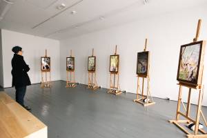 A landscape image of the Orchard Portrait installation space at Limerick City Gallery of Art. The room has white walls, black wooden floors and a wooden bench in the middle of the room. In the room are six screens on wooden easels. Each screen shows a video of a senior performer against a different branch